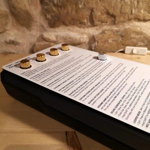 May include: A black electronic music device with gold knobs and a silver knob. The device has a white paper overlay with text and is set on a wooden surface. The device is angled towards the viewer.