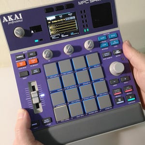 May include: A purple and grey Akai MPC Sample music production centre. The device has numerous buttons, pads, and knobs, with a screen displaying waveforms. The text "MPC SAMPLE" and "AKAI professional" are visible.