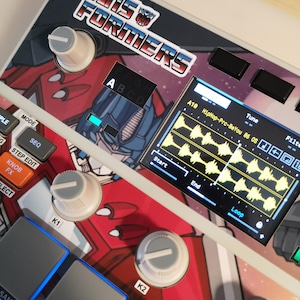 May include: A close-up of a white and red electronic music production device with the text "TRANSFORMERS". The device has knobs, buttons, and a screen displaying a waveform. The device has a Transformers graphic.