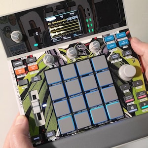 May include: A Marvel Super Heroes-themed music production device with a screen, buttons, and pads. The device features a colorful comic book design with the Marvel Comics logo and various controls for music creation. The device is held by a person.