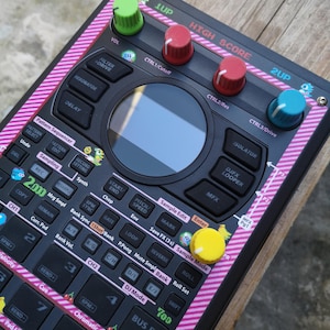 Könnte beinhalten: Ein schwarzes elektronisches Musikproduktionsgerät mit farbigen Knöpfen und Tasten. Das Gerät hat einen Bildschirm und ist mit einem rosa-weiß gestreiften Rand verziert. Der Text "HIGH SCORE" ist oben sichtbar.