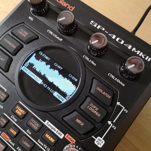 May include: Black Roland SP-404MKII sampler featuring numerous knobs, buttons, and a small screen showing a waveform. The device has a sleek design with labels for functions such as "VOL," "CTRL," and "Sample Edit."