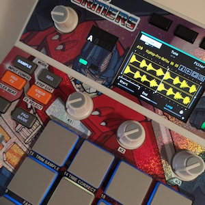 May include: A digital music production device with a Transformers theme. It features buttons, knobs, and a screen showing waveforms. The design incorporates red, blue, and silver colours, with the Transformers logo prominently displayed.