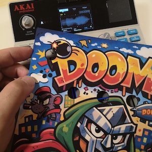 May include: A silver Akai Professional MPC Sample music production device with a screen and buttons, positioned behind a colourful custom faceplate featuring the word DOOM in large orange letters, a cartoon character in a green hood, and a bomb.
