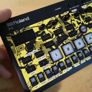 Peut inclure: Synthétiseur Roland P-6 noir et jaune avec de nombreux boutons, des molettes et un affichage numérique. L'appareil présente un motif unique et est tenu par une main. Les textes "Roland" et "P-6" sont visibles.