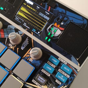May include: Electronic music production device with a black and silver design. Features a screen displaying audio waveforms, numerous buttons, knobs, and a graphic of a robot. The device has blue and white accents and is designed for music creation.