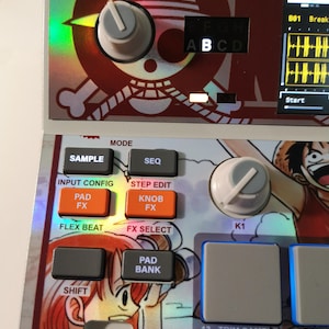 May include: Close-up of a music production device with a One Piece anime theme. The device features buttons labeled "SAMPLE", "SEQ", "PAD FX", and "KNOB FX". The design includes a skull and crossbones logo and character illustrations.