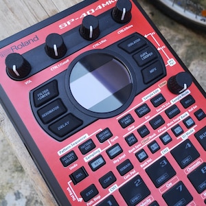 Peut inclure: Un échantillonneur Roland SP-404MKII rouge et noir avec de nombreux boutons, des molettes et un écran central. L'appareil propose des commandes pour le filtre, le délai et l'échantillonnage, avec des étiquettes comme "REC" et "Sample Edit".