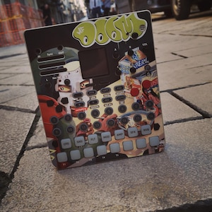 May include: A black and colorful electronic music production device with buttons, knobs, and a screen. The device has a graffiti-style design with the word "DOOM" in green. The device is sitting on a cobblestone street.