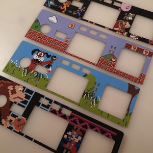 May include: Four rectangular console faceplates featuring pixel art designs from classic video games. The designs include characters like Mario, Donkey Kong, and Mega Man, with various cutouts for buttons and screens on each flat, thin panel.
