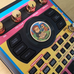 Op de afbeelding: Close-up van een kleurrijk elektronisch muziekproductieapparaat. Het apparaat heeft gouden knoppen, zwarte knoppen en een ronde sticker met stripfiguren. Het apparaat heeft tekstlabels zoals "Filter + Drive" en "Sampling".