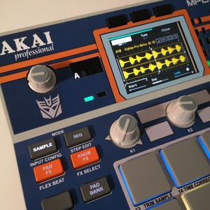 May include: A blue and orange AKAI Professional MPC series music production centre. Features include a screen displaying audio waveforms, control knobs, and various function buttons. The device has a Transformers logo.