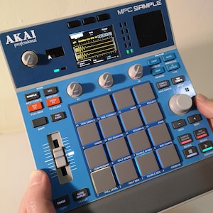 Könnte beinhalten: Ein blau-graues Akai MPC Sample Musikproduktionsgerät. Es verfügt über einen Bildschirm, Tasten, Knöpfe und einen Schieberegler. Das Gerät wird von den Händen einer Person gehalten. Der Text "MPC SAMPLE" und "AKAI professional" sind sichtbar.