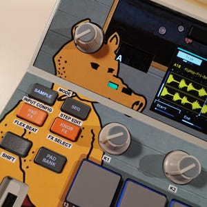May include: A music production sampler featuring a custom skin with a cartoon character illustration. The device includes grey control knobs, orange and grey buttons labelled Sample, Seq, and Pad Bank, and a digital screen displaying audio waveforms.