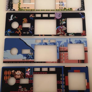 May include: Four rectangular console faceplates featuring pixel art designs. The top plate displays The Legend of Zelda text and graphics. The second plate features various video game characters. The third plate shows a blue cityscape. The bottom plate depicts a Donkey Kong game scene.