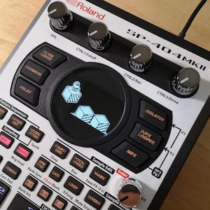May include: Roland SP-404MKII sampler with black buttons, silver knobs, and a central screen displaying a blue graphic. The device has various function buttons and is designed for music production and DJing.