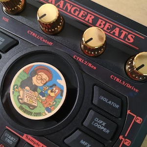 Puede incluir: Dispositivo negro de producción de música electrónica con perillas doradas y texto rojo "STRANGER BEATS". Una pegatina colorida con personajes de dibujos animados está en el centro. Botones incluyen "ISOLATOR" y "DJFX LOOPER".