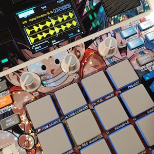 Può includere: Un campionatore per produzione musicale con una griglia di sedici pad quadrati grigio chiaro, tre manopole di controllo bianche e uno schermo digitale che mostra una forma d'onda gialla. La superficie presenta una grafica anime con stelle olografiche.