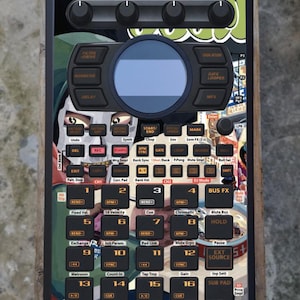 May include: Black and green electronic music production device with numerous buttons, knobs, and a central screen. Features text labels like "Filter Drive," "Delay," and "Start/End." The device has a colourful graphic design.