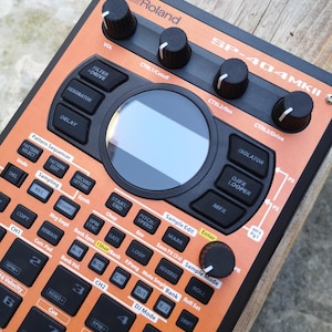 Peut inclure: Un échantillonneur Roland SP-404MKII orange et noir avec de nombreux boutons, des molettes et un écran central. L'appareil comporte des commandes pour le filtre, le délai et l'échantillonnage, avec des étiquettes telles que "Sample Edit" et "Enter".