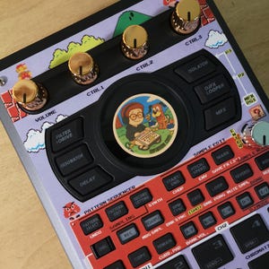 Puede incluir: Un dispositivo de producción musical personalizado con un tema de videojuegos retro. Cuenta con perillas doradas, un panel de control negro y una superposición púrpura con gráficos pixelados. Una pegatina circular con personajes de dibujos animados está en el centro.