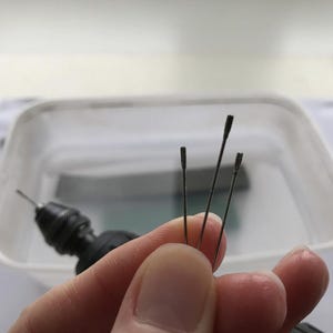 May include: Close-up of several small, dark-colored drill bits with a slightly textured surface. The bits are held in a hand, with a rotary tool visible in the background. The bits appear to be new and unused.