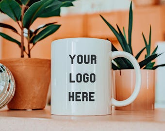 May include: White ceramic mug with the text "YOUR LOGO HERE" in black block letters. The mug is on a light pink surface, with green plants in terracotta pots in the background. A disco ball is visible on the left.