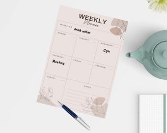 Op de afbeelding: Een weekplanner met de tekst "WEEKLY Planner" en de woorden "drink water", "Gym" en "Meeting" erop. Een blauwe en zilveren pen ligt op de planner. Een lichtgroene theepot en een klein notitieblok zijn ook zichtbaar.