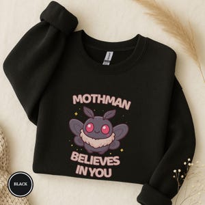 May include: Black sweatshirt with the text "Mothman Believes In You" and a cartoon moth graphic. The moth is gray with pink accents and a fluffy collar. The sweatshirt has a crew neck and long sleeves.