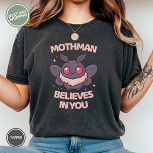 May include: Dark gray t-shirt featuring a cartoon Mothman with pink eyes and a fluffy chest. The text "Mothman Believes In You" is printed above and below the graphic in pink. The shirt has a crew neck and short sleeves.