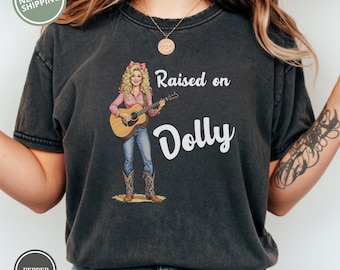 May include: Dark gray t-shirt featuring a graphic of a woman playing a guitar, with the text "Raised on Dolly." The woman is wearing a pink shirt, blue jeans, and cowboy boots. The shirt has a vintage, washed look.