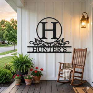 Könnte beinhalten: Schwarzes Metall-Familiennamenschild mit dem Buchstaben "H" in einem Kreis und dem Namen "HUNTER'S" darunter. Das Schild ist an einer weißen Wand über einer Veranda mit einem Schaukelstuhl und Topfpflanzen angebracht.