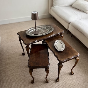 Peut inclure: Un ensemble de trois tables gigognes antiques en bois avec des pieds incurvés et un fini foncé et brillant. Les tables présentent un veinage du bois. Un plateau argenté et une petite lampe marron sont posés sur la plus grande table.