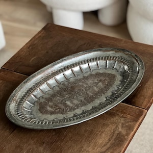 Può includere: Un vassoio ovale in metallo argentato con un bordo decorativo e un design centrale dettagliato. Il vassoio è posto su una superficie di legno scuro, mettendo in risalto la sua estetica vintage. Le dimensioni del vassoio non sono visibili.