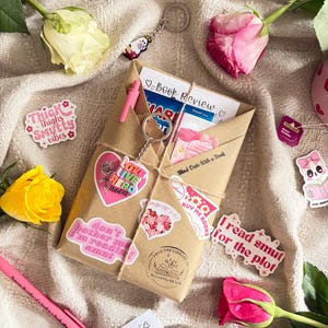 May include: A brown paper-wrapped book package tied with twine, featuring a pink pen, a heart-shaped keychain, and various stickers with text like 'Love Live Read Addict' and 'I read smut for the plot'. Includes book review cards and fresh roses.