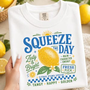May include: A white crew neck t-shirt featuring a graphic print with a yellow lemon, green leaves, and blue text that reads Squeeze the Day, Zesty & Bright, Main Character Energy, Fresh Pick, Citrus Stand Special, Tangy, Happy, and Golden.