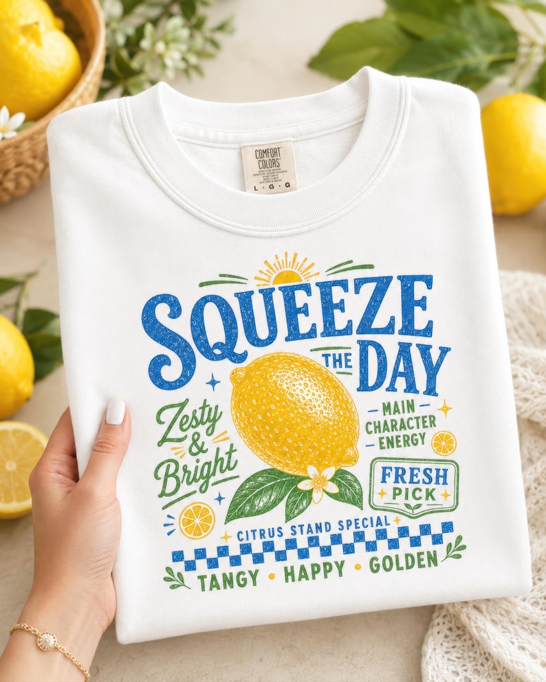May include: A white crew neck t-shirt featuring a graphic print with a yellow lemon, green leaves, and blue text that reads Squeeze the Day, Zesty & Bright, Main Character Energy, Fresh Pick, Citrus Stand Special, Tangy, Happy, and Golden.