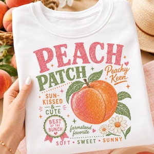 May include: A white sweatshirt featuring a colorful graphic print of a peach with green leaves and daisies. The text reads PEACH PATCH, Peachy Keen, SUN-KISSED & CUTE, BEST OF THE BUNCH, farmstand favorite, and SOFT SWEET SUNNY in various fonts and colors.