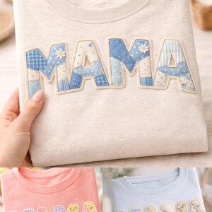 以下が含まれることがあります： 前面に「MAMA」の文字がアップリケされたベージュのスウェットシャツ。文字は、さまざまな青と白の色合いのパッチワーク生地で作られており、花柄や模様のディテールが施されています。「A」の文字にはデイジーが飾られています。