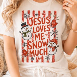 May include: A white short-sleeved t-shirt featuring a red vertical striped background with the text "JESUS LOVES ME SNOW MUCH" in red letters. The design includes two snowmen, red mittens, and black and white snowflakes scattered throughout.