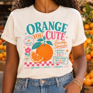 May include: A cream-colored t-shirt featuring a graphic of a whole orange and a slice with green leaves. The text reads: ORANGE YOU CUTE, MARKET BEST, FRESHLY SQUEEZED, SUNNY DAYS ONLY, GROVE-GROWN GOODNESS, BRIGHT, JUICY, CHEERY, with a pink checkered border.