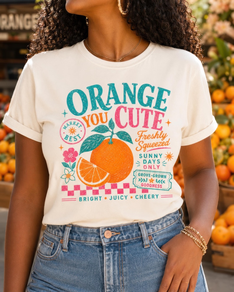 May include: A cream-colored t-shirt featuring a graphic of a whole orange and a slice with green leaves. The text reads: ORANGE YOU CUTE, MARKET BEST, FRESHLY SQUEEZED, SUNNY DAYS ONLY, GROVE-GROWN GOODNESS, BRIGHT, JUICY, CHEERY, with a pink checkered border.