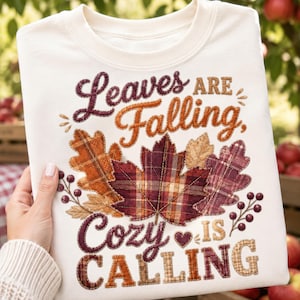 May include: A cream-colored sweatshirt featuring a graphic design with the text "Leaves ARE Falling, Cozy IS CALLING" in brown and maroon. The design includes a central arrangement of autumn leaves in plaid patterns of orange, red, and brown.