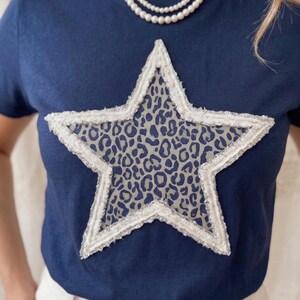 May include: Navy blue t-shirt featuring a large star design. The star has a white, textured border with silver accents and a leopard print center. The shirt is a classic crew neck style.