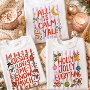 May include: Three white crewneck sweatshirts featuring festive holiday graphics. One displays "ALL IS CALM, Y'ALL" with a moon and candles. Another shows "JESUS LOVES ME SNOW MUCH" with snowmen. The third reads "HOLLY JOLLY EVERYTHING" with ornaments.