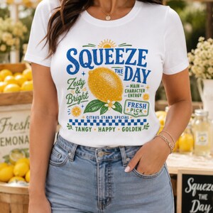 May include: A white short-sleeve t-shirt featuring a graphic print of a yellow lemon with green leaves and the text SQUEEZE THE DAY, Zesty & Bright, Main Character Energy, Fresh Pick, Citrus Stand Special, Tangy, Happy, Golden in blue and green.