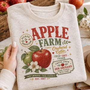 May include: A heathered beige crewneck sweatshirt featuring a graphic print with the text APPLE FARM, Orchard Cutie, and Crisp Cozy Charm. The design includes illustrations of a red apple, a green apple, apple blossoms, and a stamp reading Picked to Perfection.