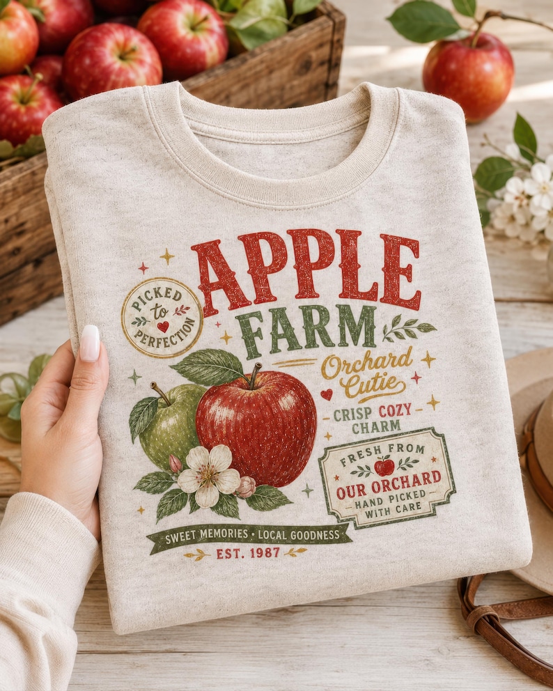 May include: A heathered beige crewneck sweatshirt featuring a graphic print with the text APPLE FARM, Orchard Cutie, and Crisp Cozy Charm. The design includes illustrations of a red apple, a green apple, apple blossoms, and a stamp reading Picked to Perfection.