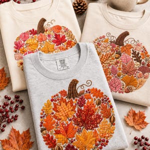 May include: Three crewneck sweatshirts in cream, light beige, and heather grey, each featuring a large embroidered pumpkin graphic composed of various patterned autumn leaves, acorns, and berries. The grey sweatshirt has a Comfort Colors label.