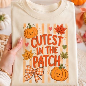 May include: A cream-colored t-shirt featuring a graphic print with vertical orange stripes, two smiling orange pumpkins, autumn leaves, small hearts, and a checkered bow. The text reads "CUTEST IN THE PATCH" in bold orange letters.